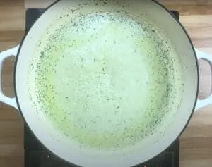 Cooking creamy sauce in a white pot on a stovetop with herbs sprinkled on top.
